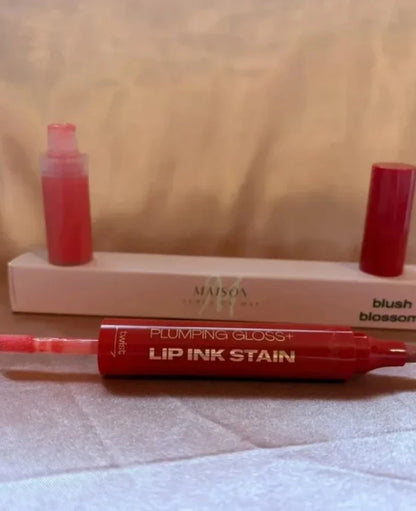 Maison Cosmetics Plumping Gloss+ Lip Ink Stain in 'blush blossom' shade, showing the red applicator pen in the foreground and the product bottle and cap on a beige box in the background.