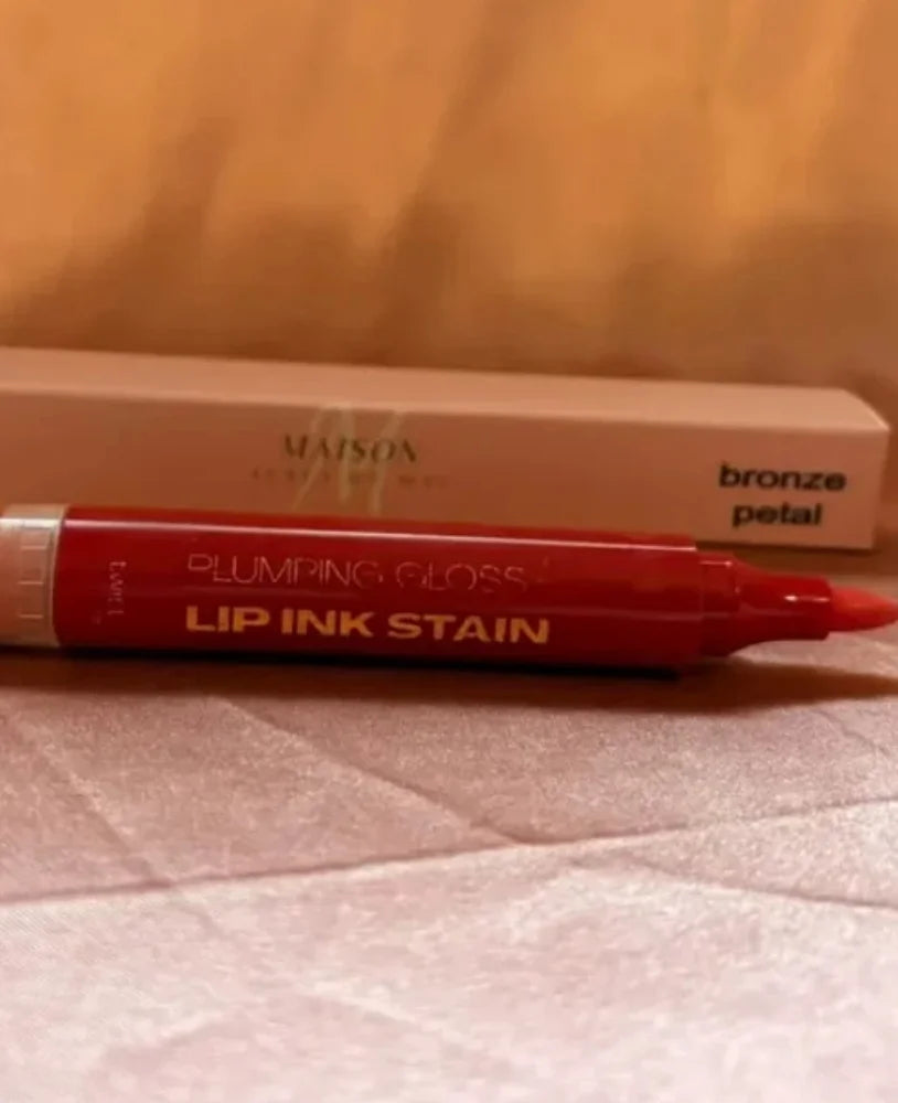A red Maison Beauty Plumping Gloss Lip Ink Stain pen in the shade bronze petal, shown in front of its beige cardboard packaging box on a textured light surface.