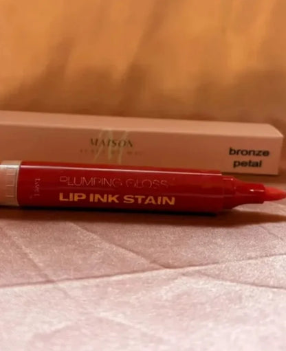 A red Maison Beauty Plumping Gloss Lip Ink Stain pen in the shade bronze petal, shown in front of its beige cardboard packaging box on a textured light surface.