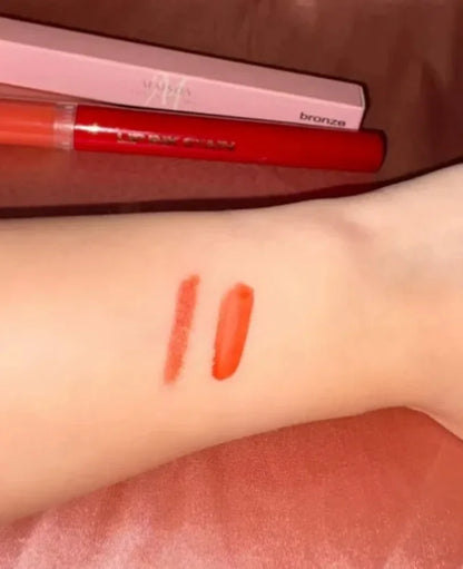A close-up of a person's forearm with two orange-red lipstick swatches applied, with a pink and red lipstick tube and box visible in the background.