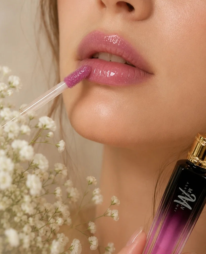 Close-up of a model applying a glossy mauve-pink lip product with a clear applicator wand. The product bottle, which has a black-to-purple gradient and a gold cap, is visible in the lower right corner, with white baby's breath flowers on the left.