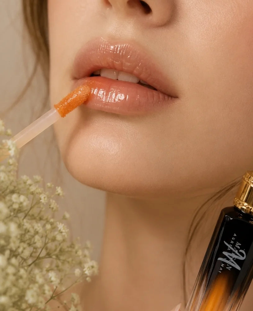 Close-up of a woman's lips wearing a glossy peachy-orange lip product, with the applicator resting on her upper lip. A small bottle with a gold cap and white script logo is partially visible in the lower right, and dried white flowers appear on the left.