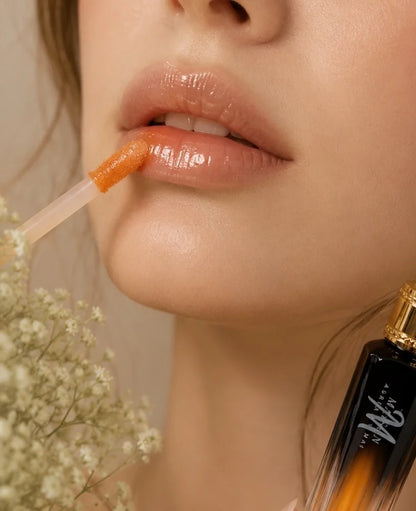 Close-up of a woman's lips wearing a glossy peachy-orange lip product, with the applicator resting on her upper lip. A small bottle with a gold cap and white script logo is partially visible in the lower right, and dried white flowers appear on the left.