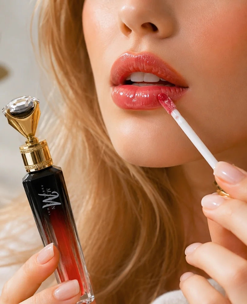 A close-up lifestyle shot of a woman applying a glossy pink-red lip gloss with a doe-foot applicator, while holding a black and red gradient lip gloss bottle with a gold cap.
