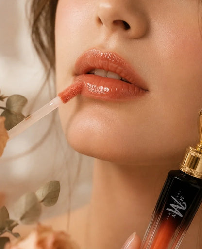 Close-up of a model applying a glossy peachy nude lip product with a doe-foot applicator, with the branded bottle partially visible in the lower right corner.