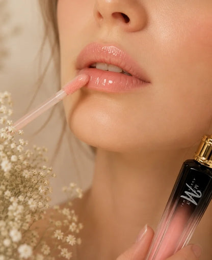 Close-up of a woman applying a glossy pink lip product with a doe-foot applicator, holding a black-to-pink gradient tube with a gold cap.